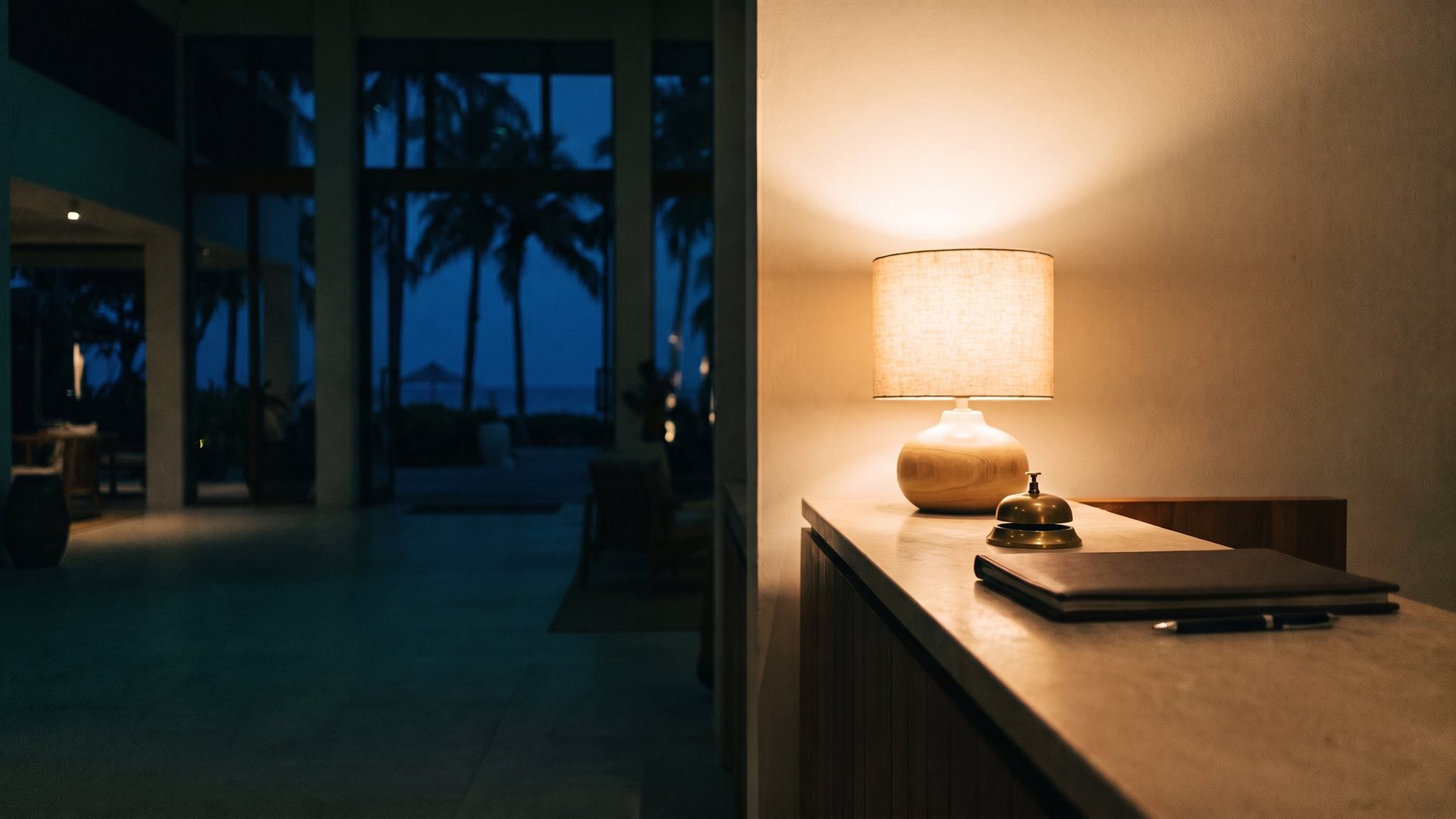 An empty resort front desk at night lit by one warm lamp, a brass service bell on the counter and palms through the lobby windows